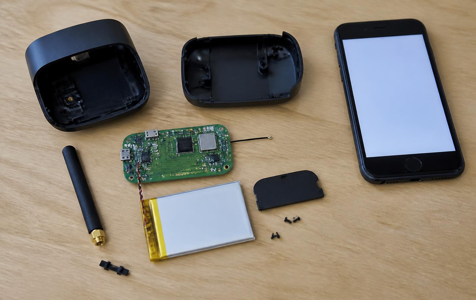 Disassembled connected device showing PCB, lithium battery, antenna and plastic housing next to a phone