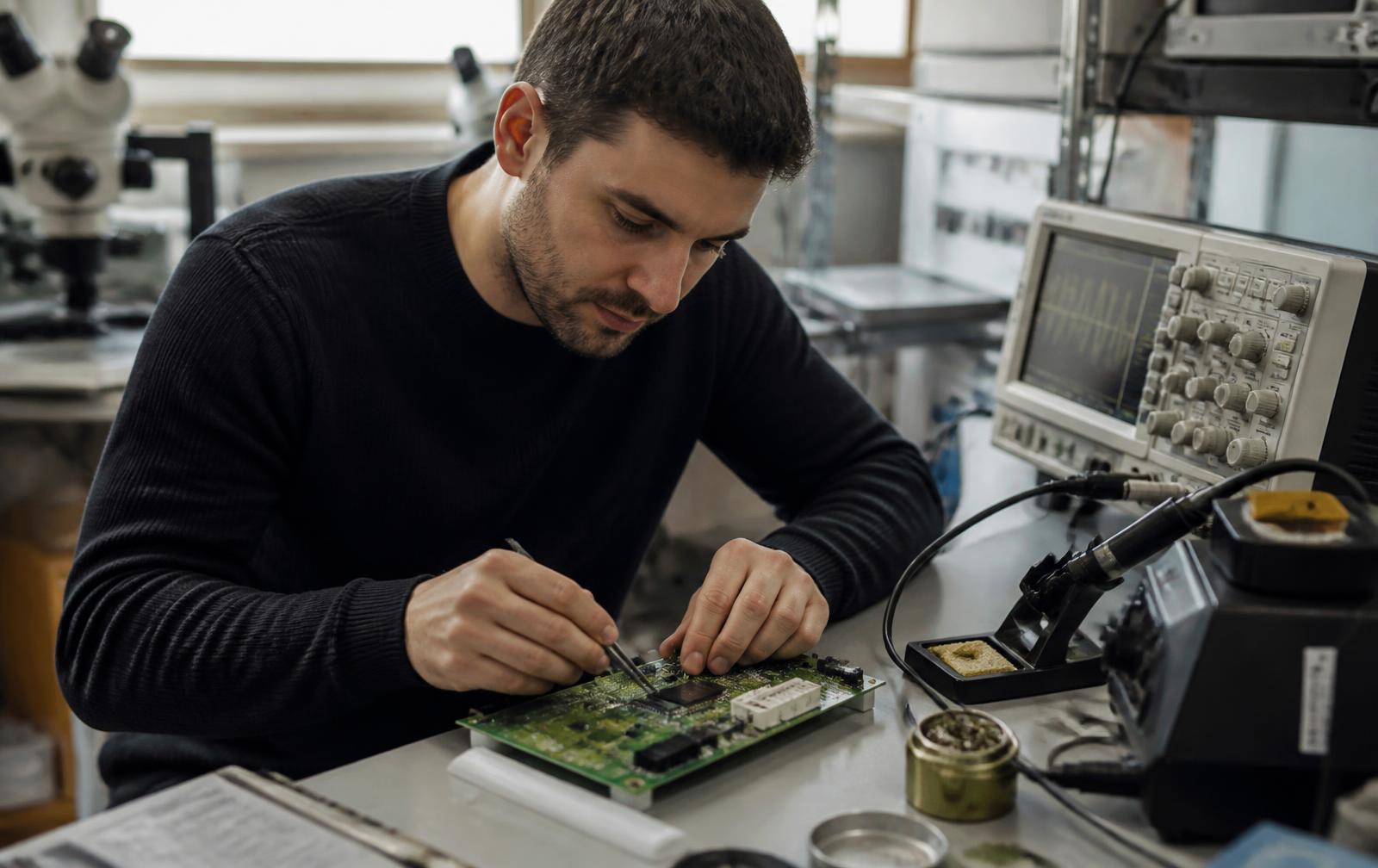 Electronics engineer probing a green printed circuit board prototype at a lab bench with an oscilloscope and soldering station