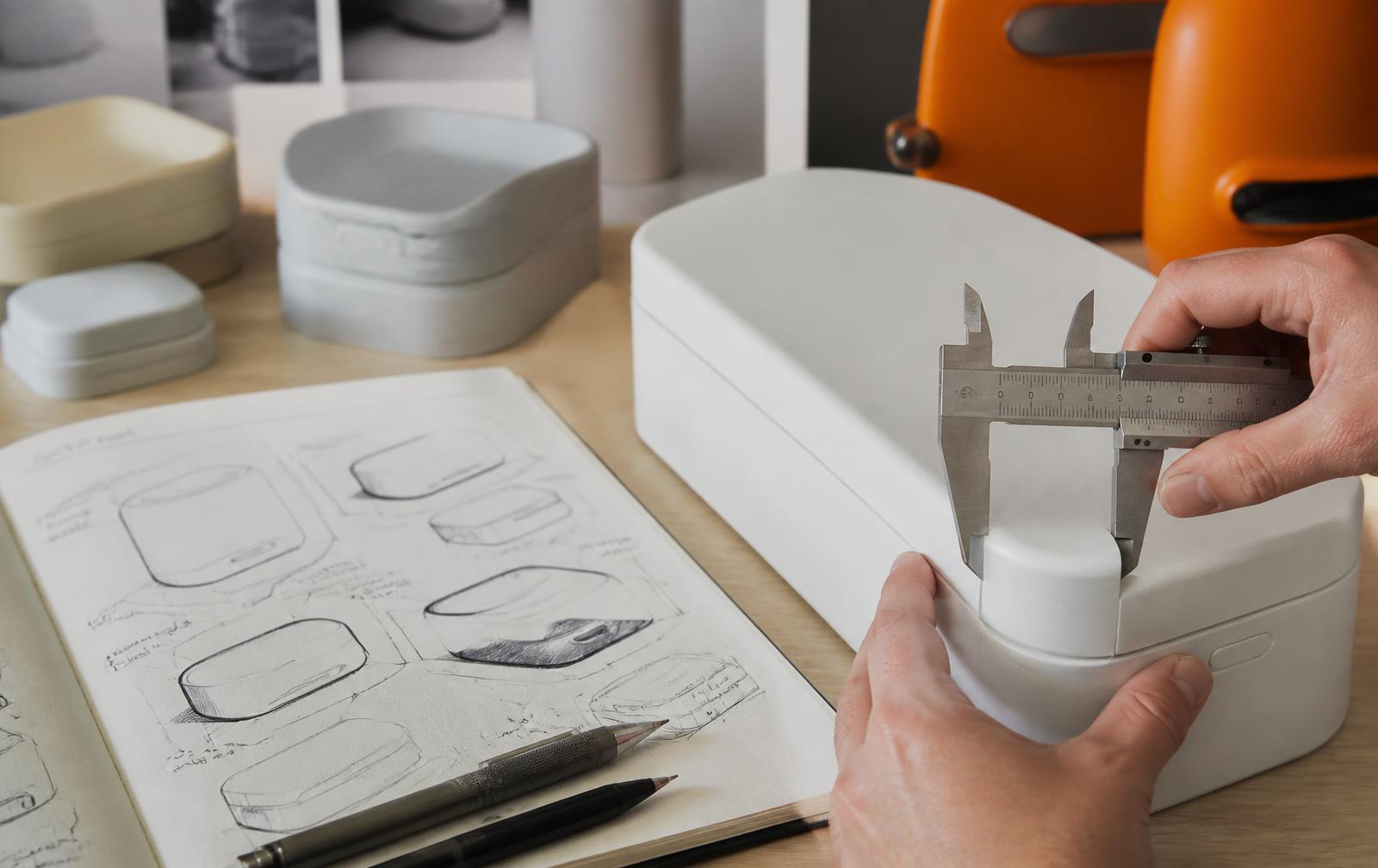 Designer measuring the edge radius of a white product prototype with calipers beside foam models and concept sketches