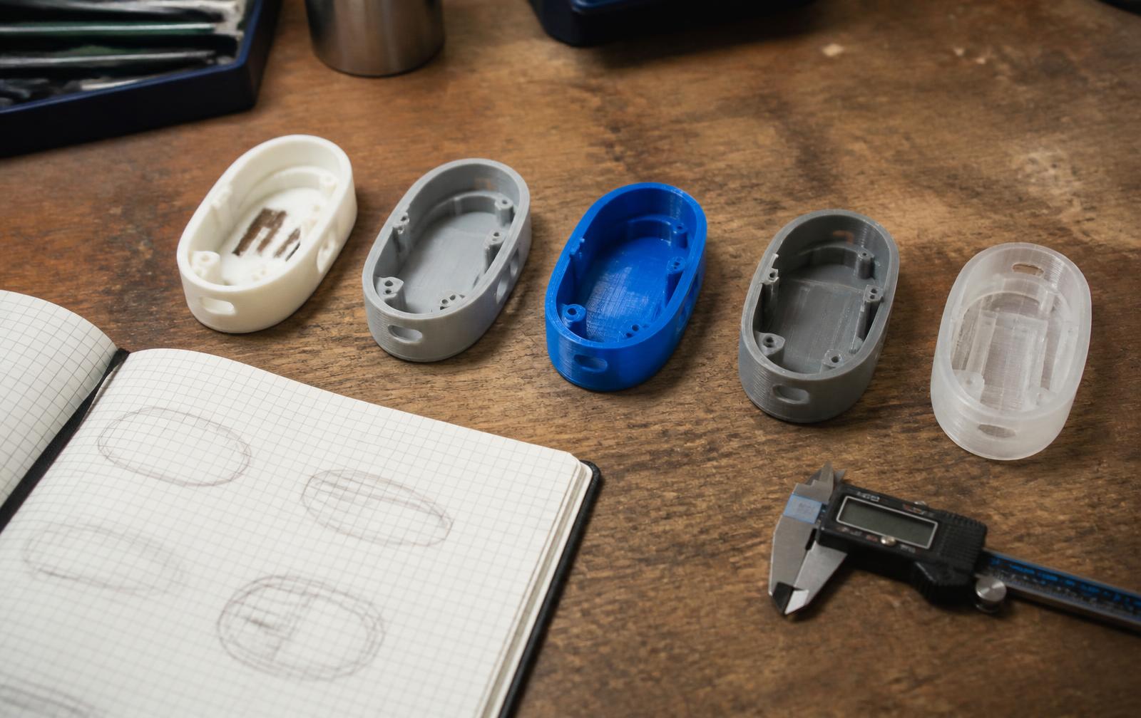 Five 3D printed versions of the same small oval plastic housing on a workbench with calipers and a sketchbook