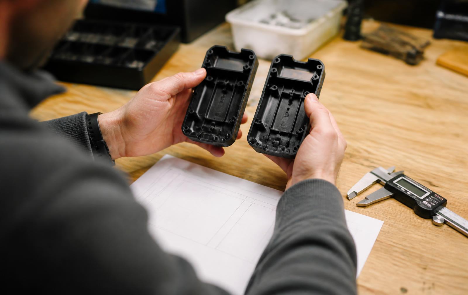 Engineer comparing two black plastic enclosure prototypes at a workbench with calipers and a spec sheet