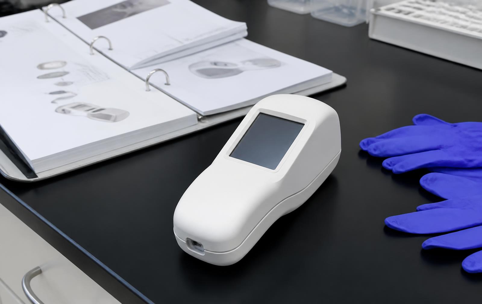 White handheld medical diagnostic device prototype on a lab bench beside a design history binder and gloves